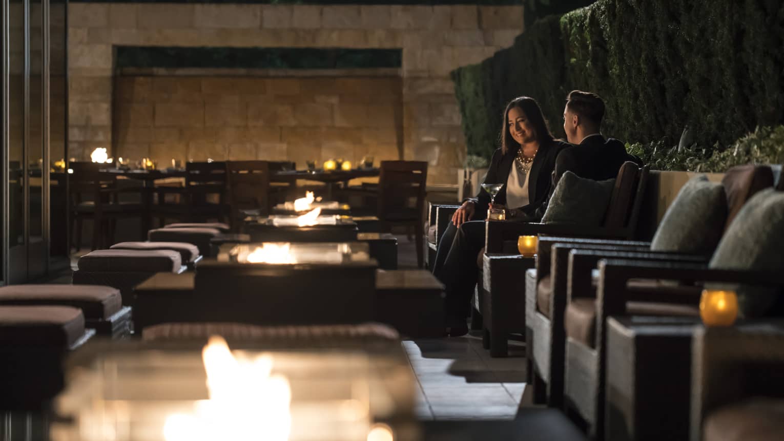 Two people in business attire sit in candle-lit cocktail lounge chairs with martinis