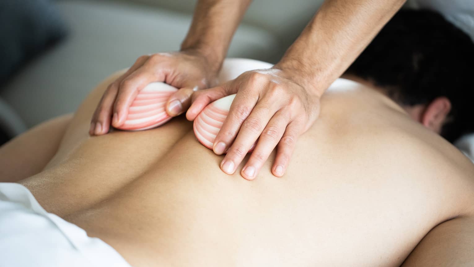 A masseuse's hands hold two white, pink-striped lava shells, running over the bare back of a man lying face down on a massage table