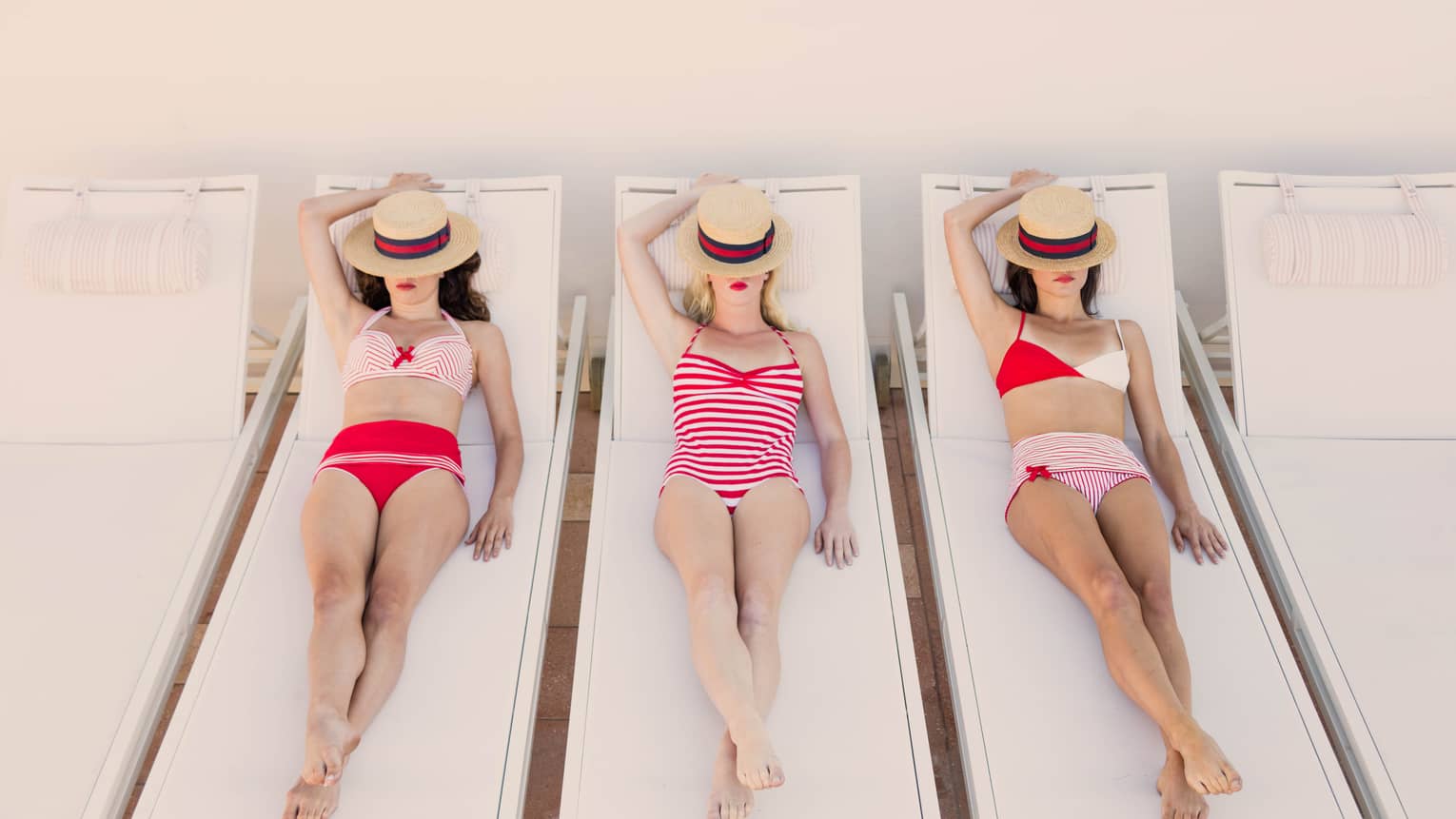 Three women in retro-style red striped bikinis lounge on pool chairs, straw fedoras over faces 