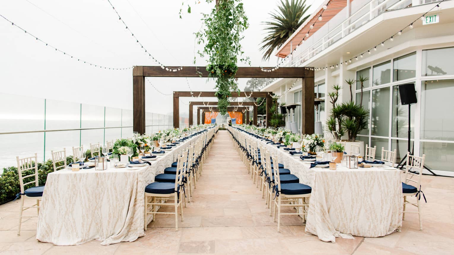 Two long dining tables on patio under strings of lights, vines, wood arches