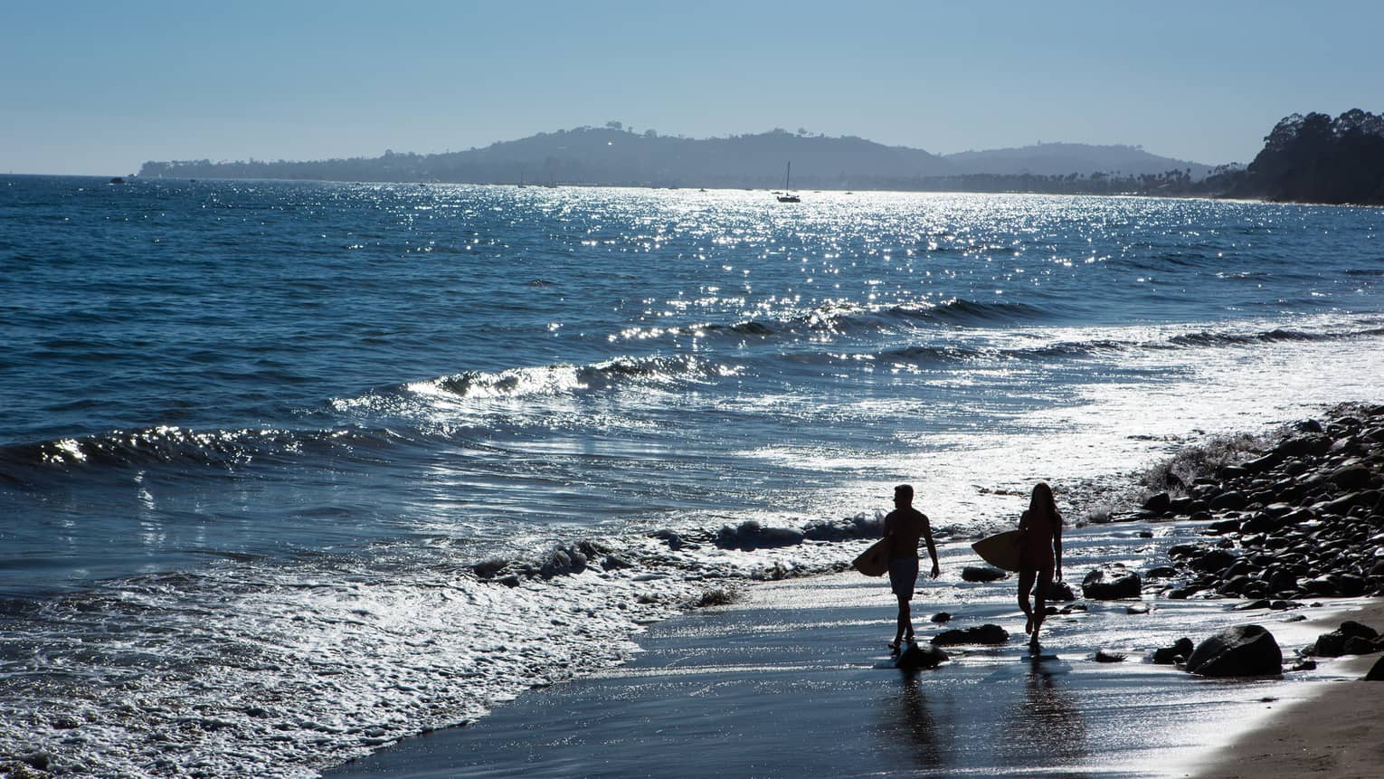 Silhouettes of two people carrying surfboards along beach shoreline