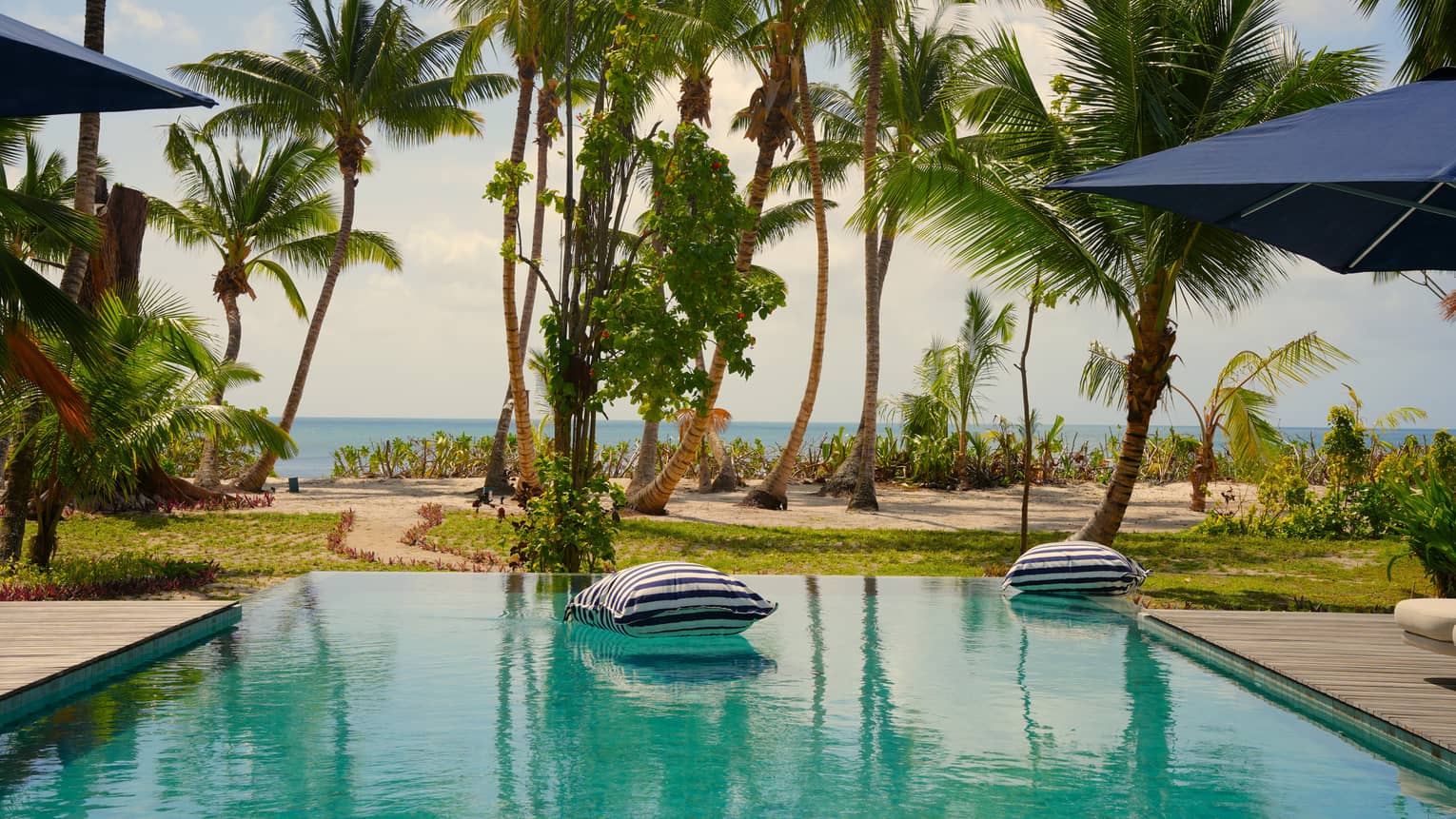 Large floating pillows float on private pool of a tropical residence, with the beach and ocean in the distance