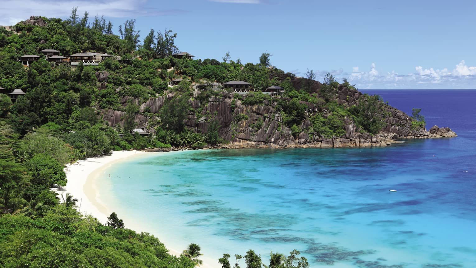 Looking down at bay with white sand beach, blue-green water, resort buildings along lush cliff, rocks