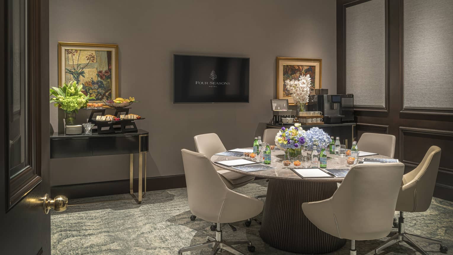 Round marble table with six chairs surrounding, wall-mounted TV at front of room, snack display visible off to the left