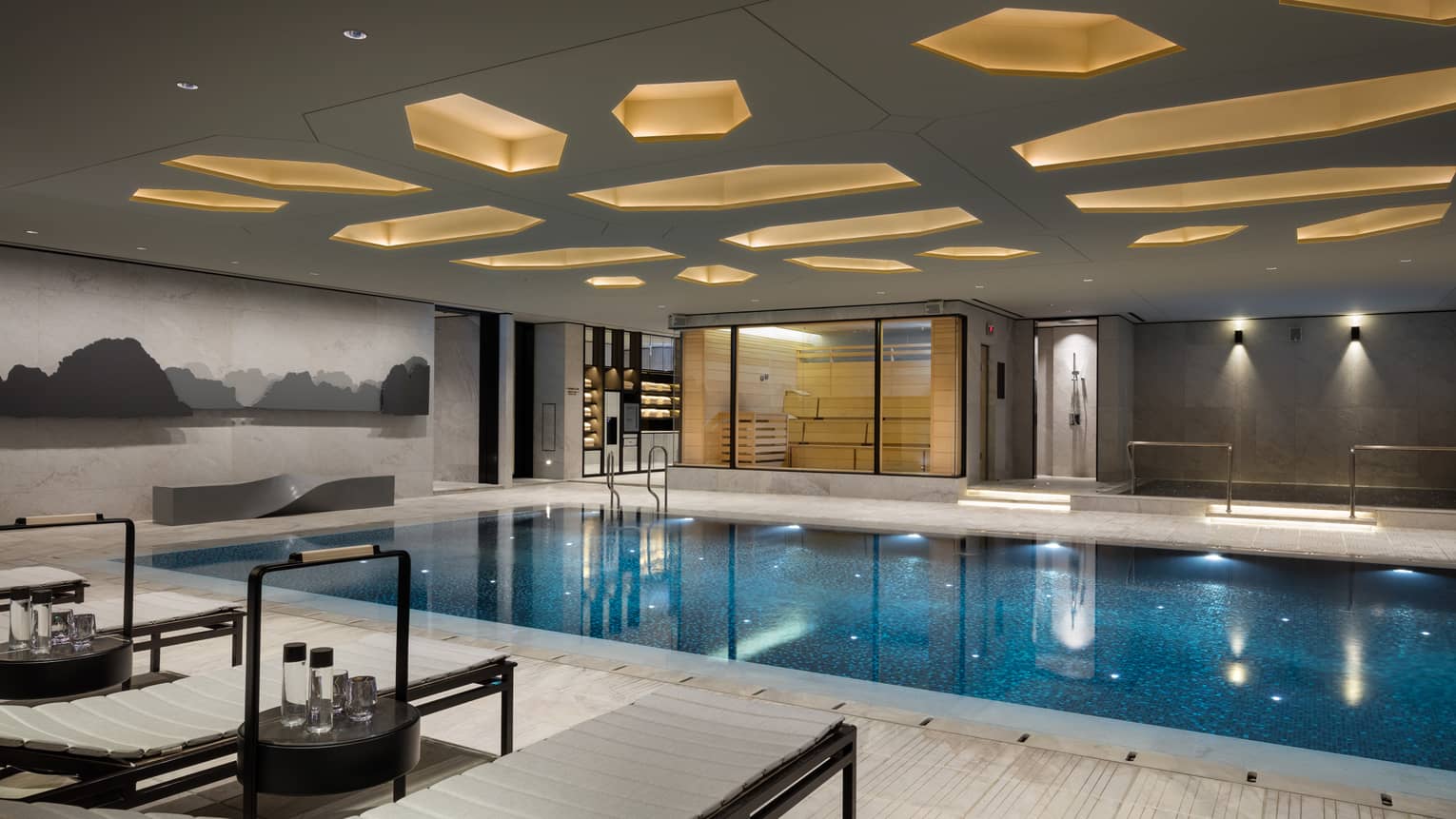 Lounge chairs, tables with water on deck by indoor swimming pool under ceiling with decorative recessed lights