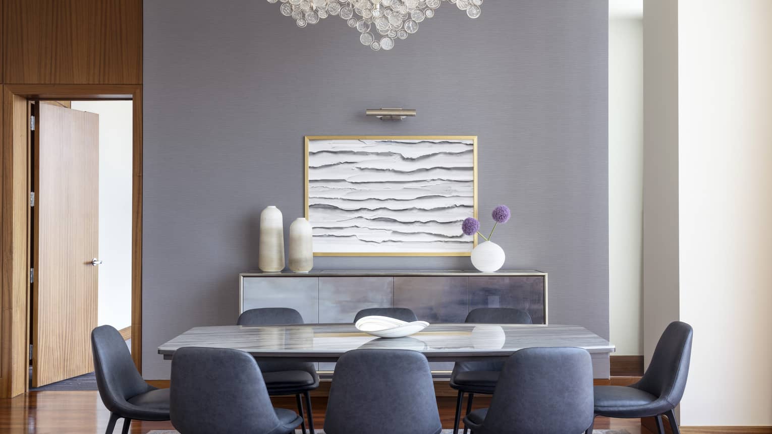 Luxury suite dining area with a marble table, dark blue chairs, a decorative sideboard, abstract wall art and a glass bubble chandelier