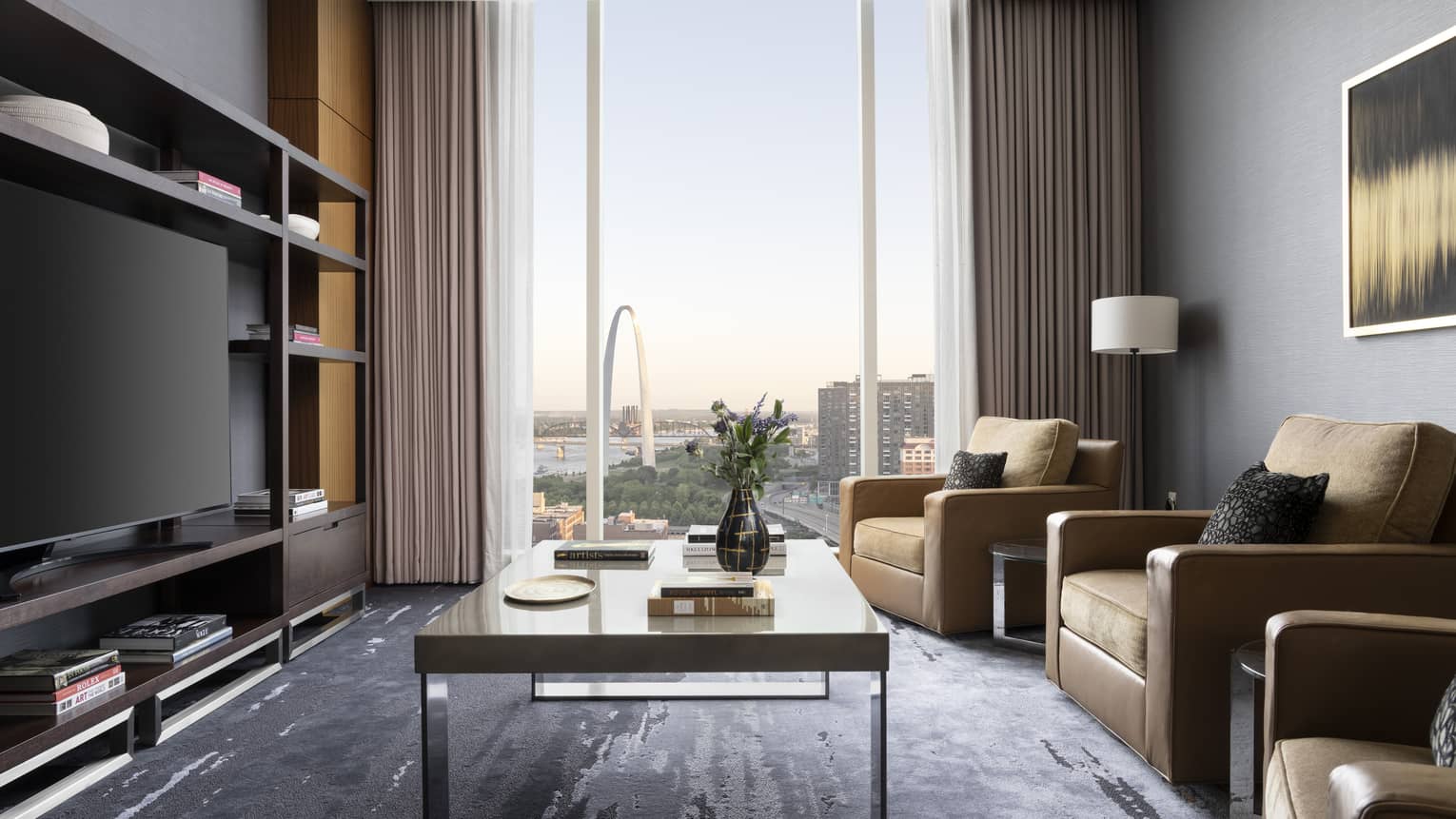Luxury suite media room with large windows offering a view of the Gateway Arch, TV, three armchairs and a coffee table with books and a vase with flowers