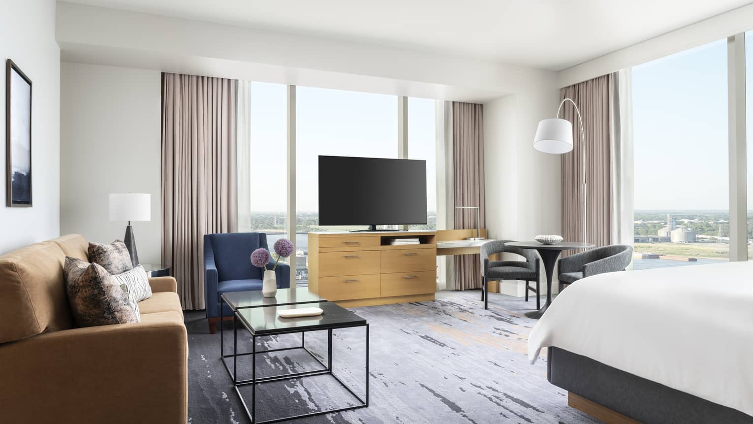 Hotel room with a beige sofa, blue armchair, coffee tables, TV on a wooden stand, table with chairs and large windows with a river view