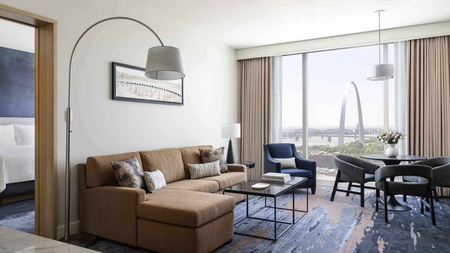 Living area with a beige sofa, coffee table, table with chairs, blue armchair, and floor-to-ceiling windows with a view of the Gateway Arch and river