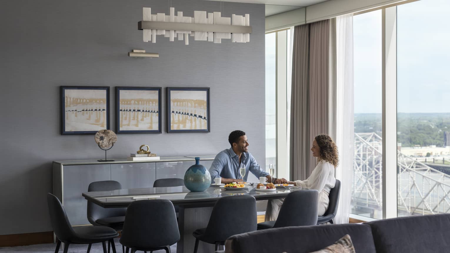 Man and woman sit at dining table in hotel suite's living room, there pieces of artwork on the wall, wall of windows