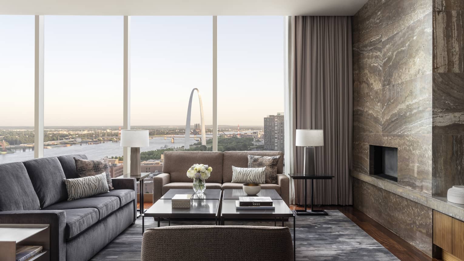 Luxury suite living room with large windows offering a view of the Gateway Arch and river, gray and beige sofa, coffee tables and a stone accent wall