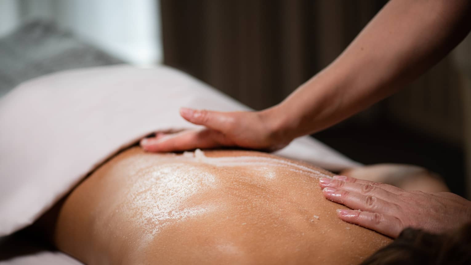 A masseuse spreads a cream across a woman's back as she lies on a massage table.