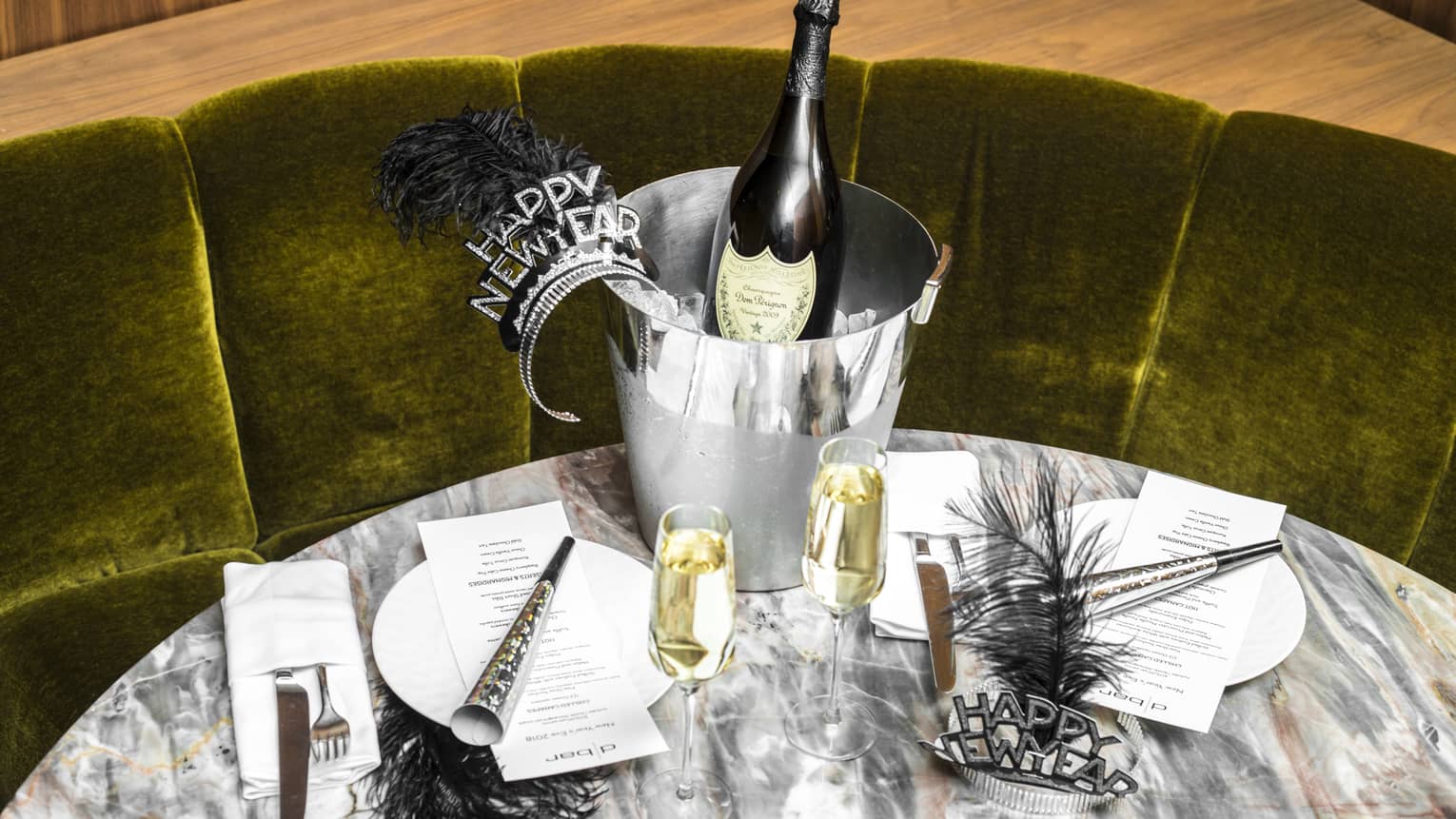 Green velvet banquette and a marble round table are set with New Year's Eve decorations, including two glasses of champagne, black feather headbands, cone-shaped noisemakers, a bottle of champagne sitting in a silver ice bucket and two sets of plates and cutlery