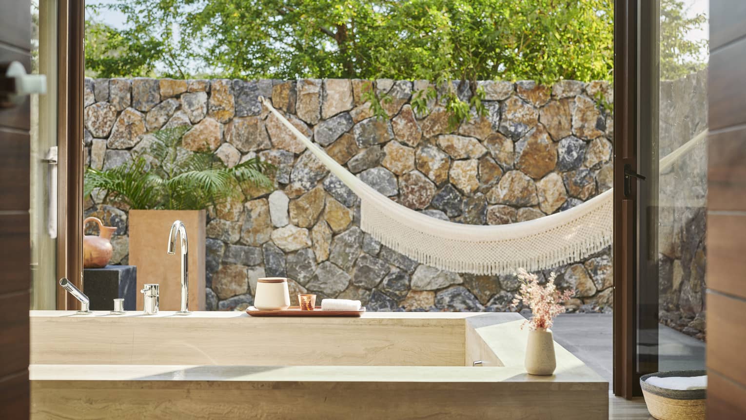 A bathroom with a large bath and large window looking outside at a hammock and stone wall.