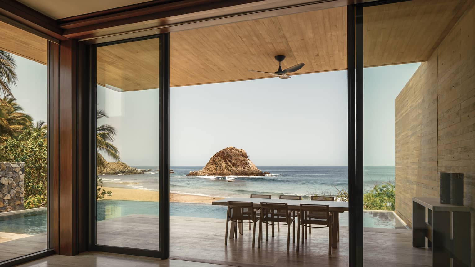 A serene ocean view through large glass doors, featuring a dining table on a patio. A small rocky island sits in the distance, framed by palm trees and a private pool.