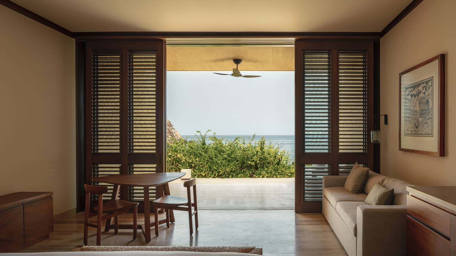 Cozy bedroom with wooden shutters, a sofa and a small dining table. The room opens to a private terrace with ocean views, framed by greenery and a ceiling fan.