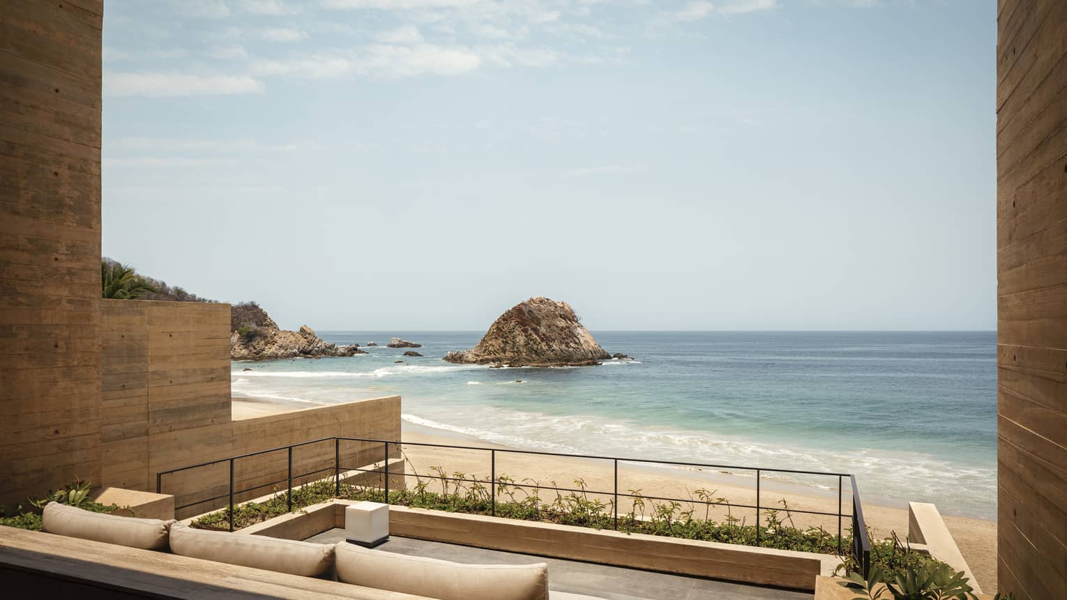 A relaxing beachfront patio with comfortable seating, offering unobstructed views of a sandy beach and rocky islands against the calm blue ocean under a clear sky