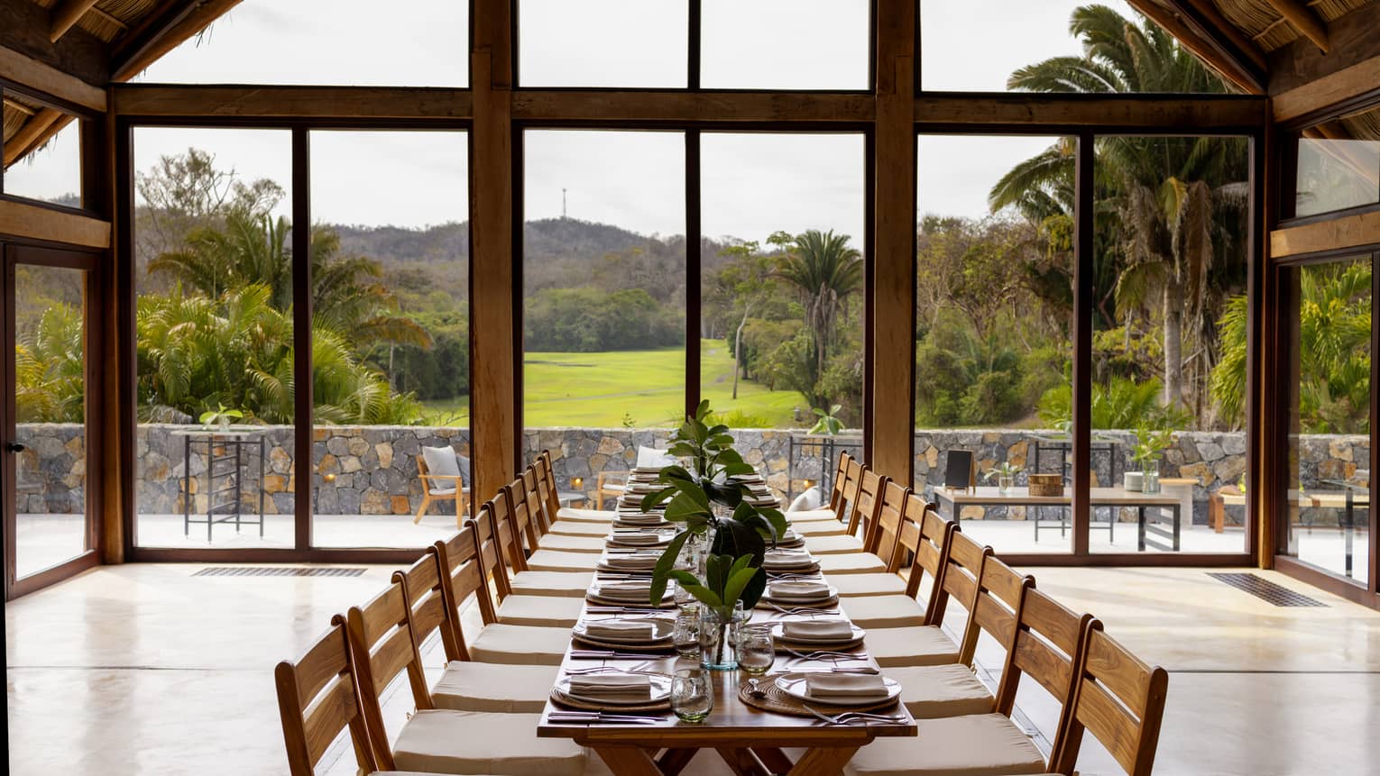 Room with floor-to-ceiling windows and a view of greenery and the mountains, with a rectangular table and place settings.