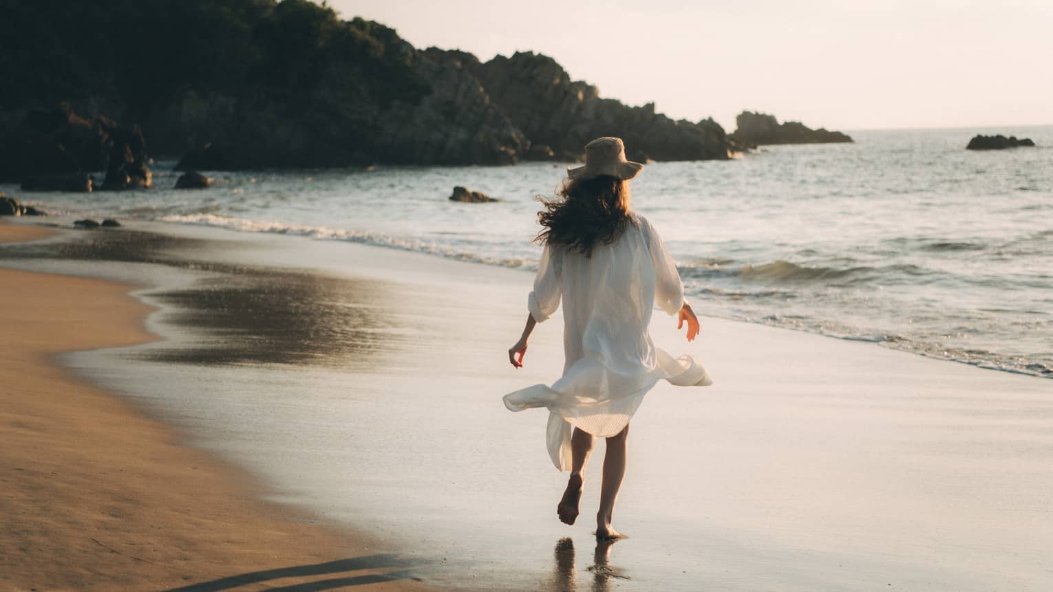 A woman in a long white shirt walking along a beach.