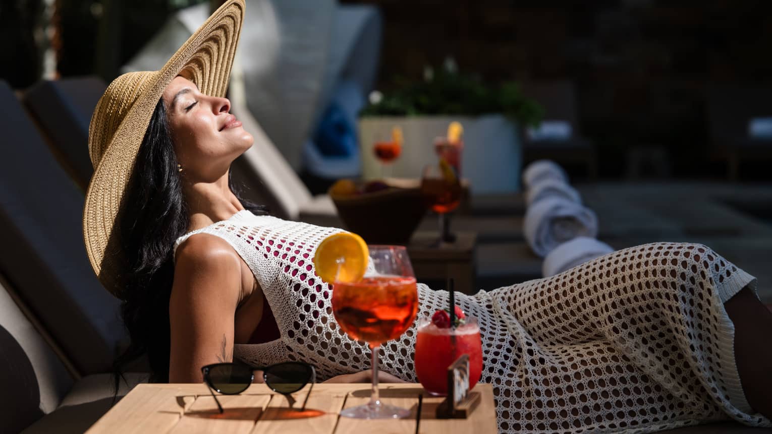 A guest laying in a lounge chair in the sunshine with their sunglasses and drink on a table beside them