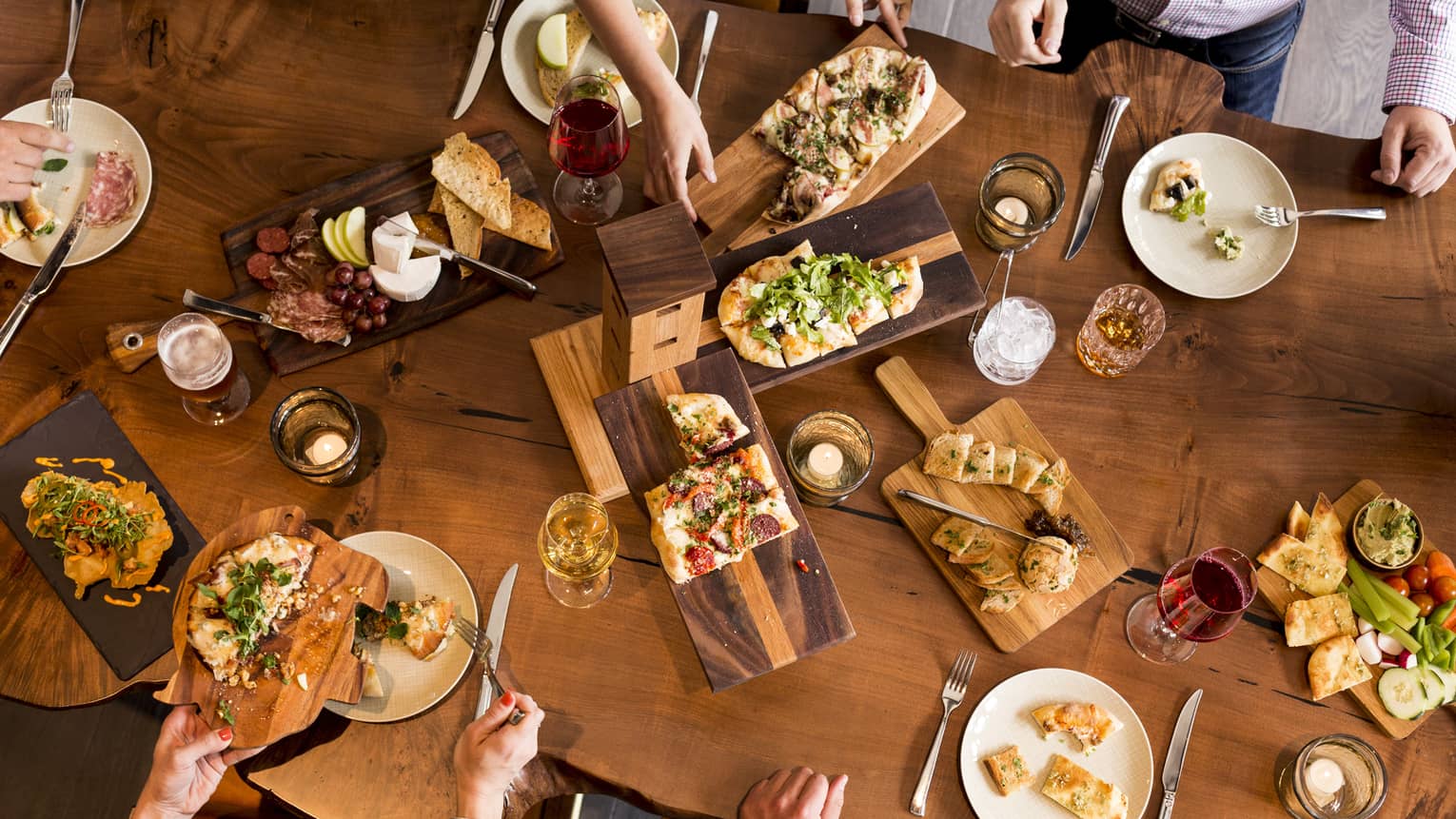 Aerial view of people dining around rustic wood table with platters of flatbreads, appetizers