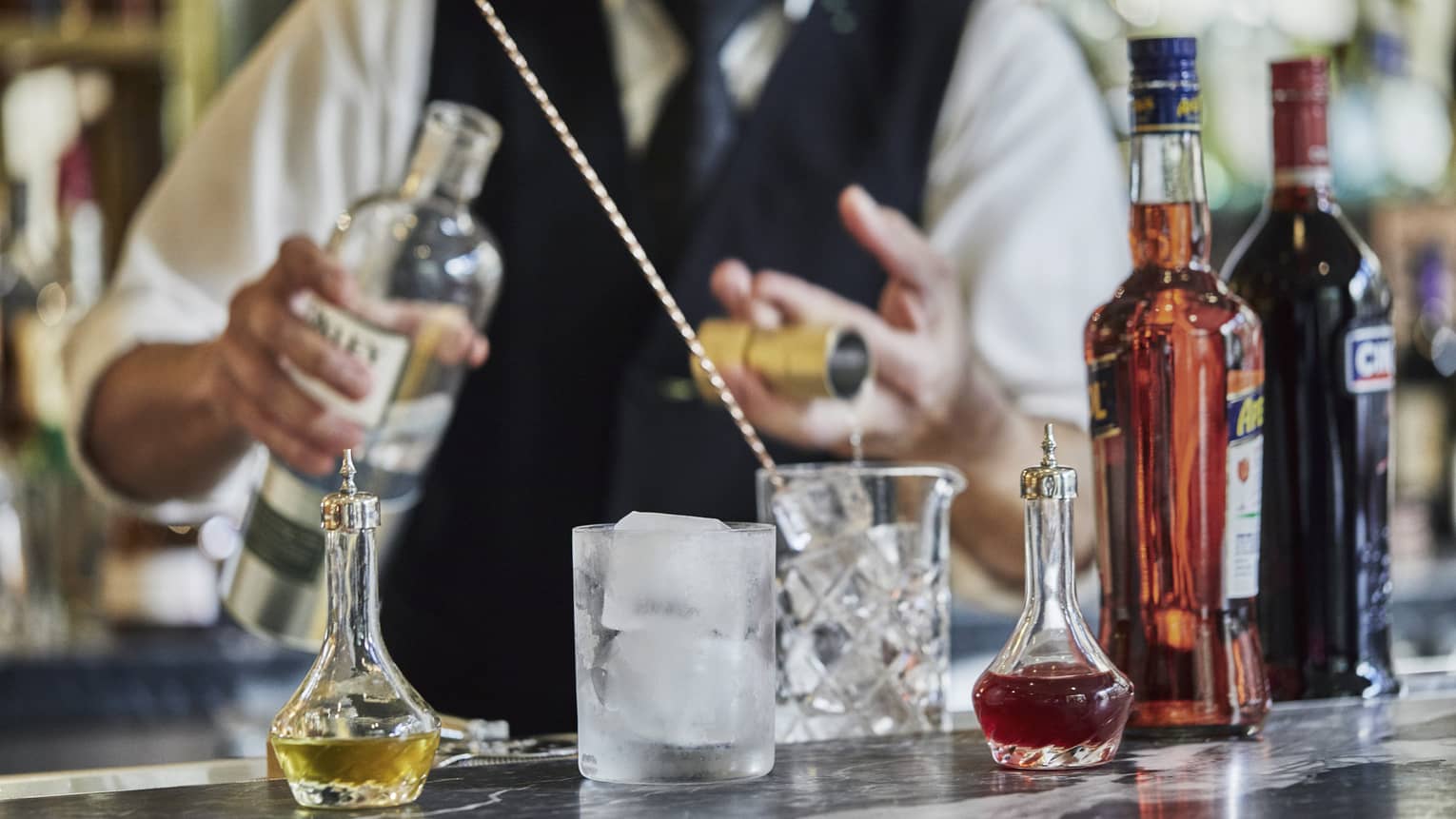 Mixologist pouring liquor into crystal cocktail shaker, glass with large ice cubes on bar
