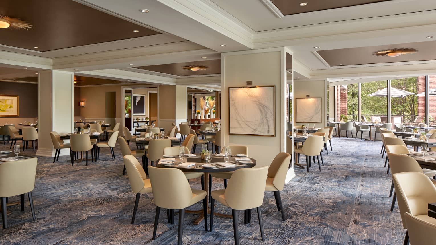A well lit restaurant with beige chairs and round tables and large open windows.