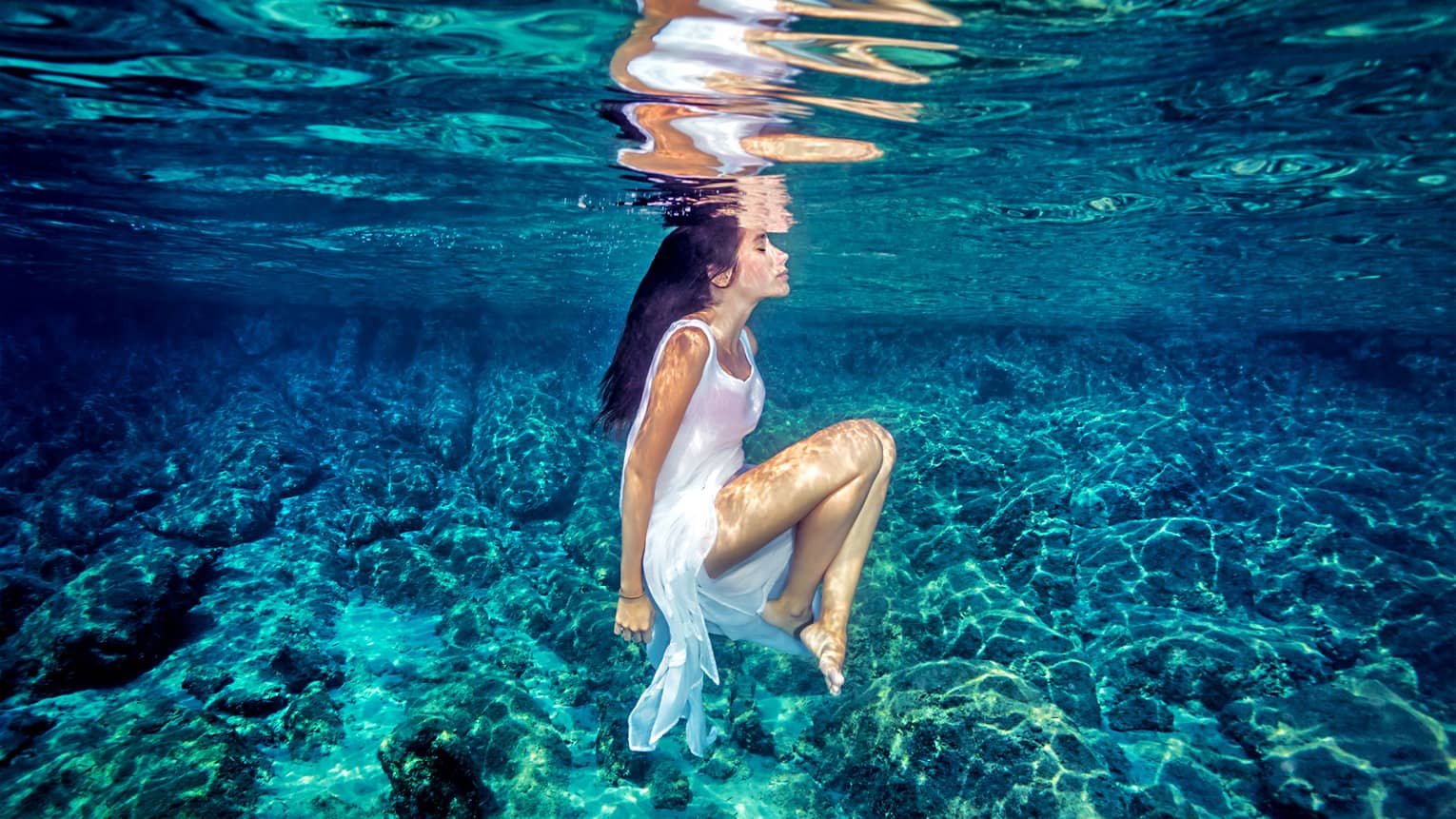 Person in a white dress floating underwater over a clear, rocky seabed, illuminated by sunlight