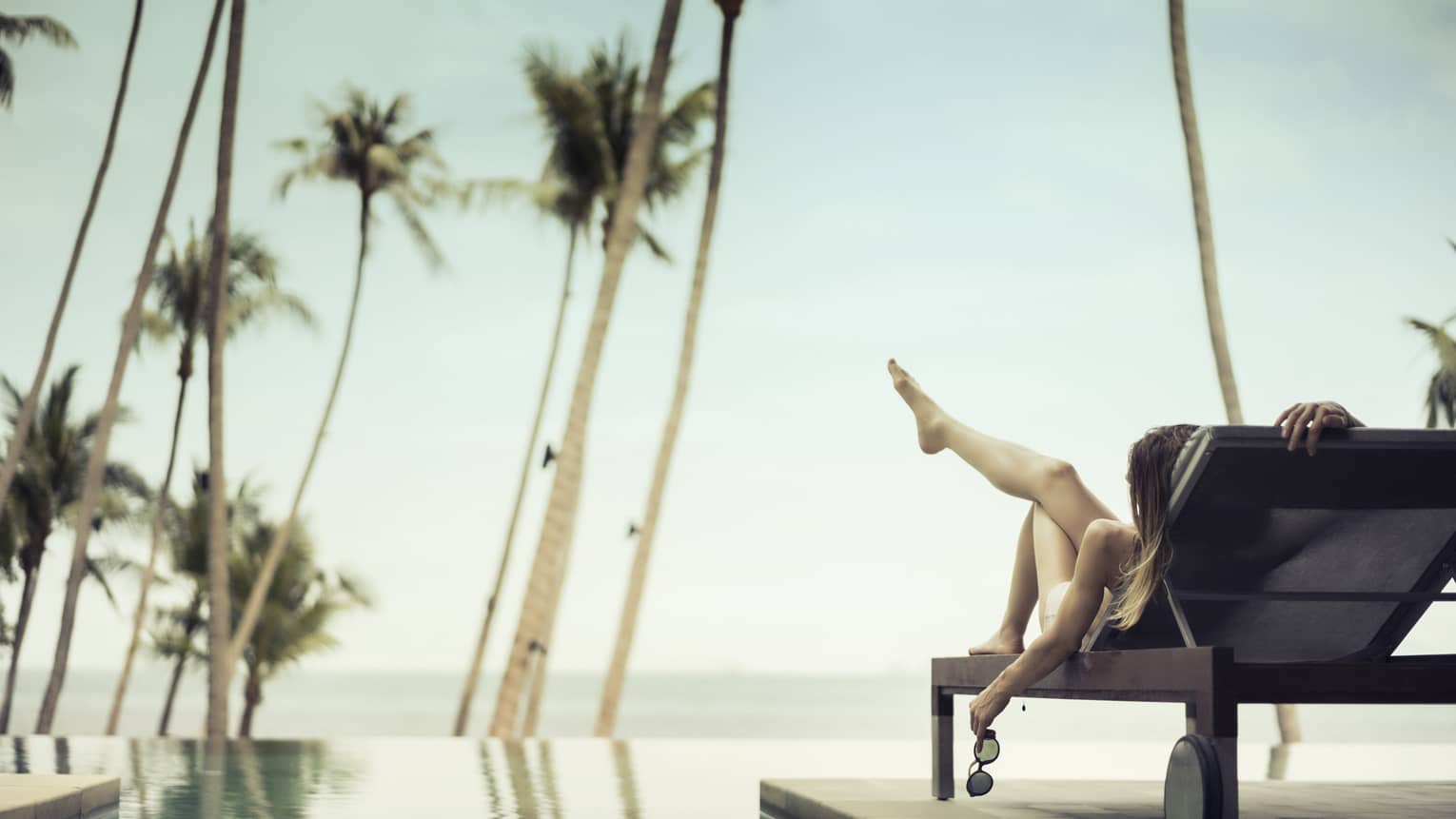 Back view of woman lounging in patio chair with leg in the air, sunglasses in hand, in front of swimming pool