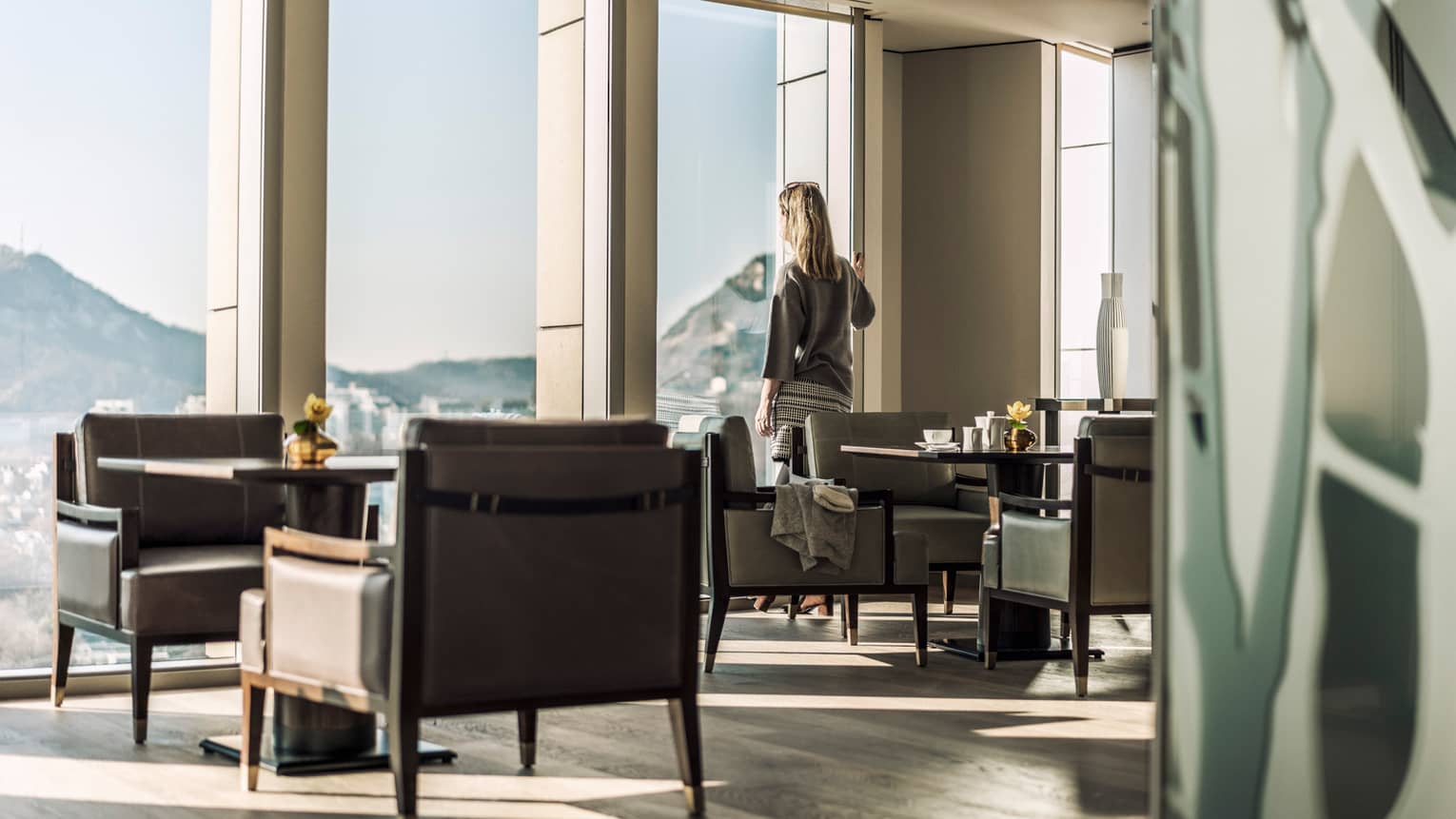 Woman stands at sunny floor-to-ceiling window and looks out at view, near lounge tables, chairs