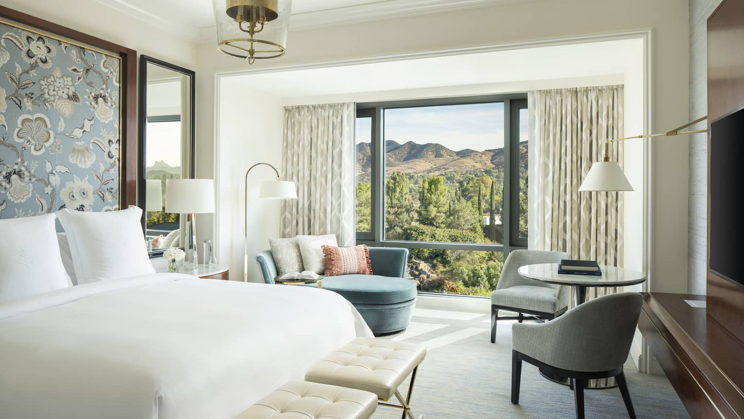 Guest room with a King bed and floral wallpaper, a blue chaise lounge, small round table with two grey chairs, and a window