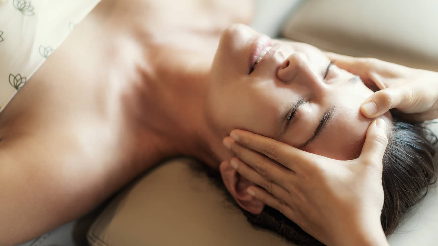 Spa facial, two hands rest on woman's forehead as she closes her eyes, lays under sheet on treatment table