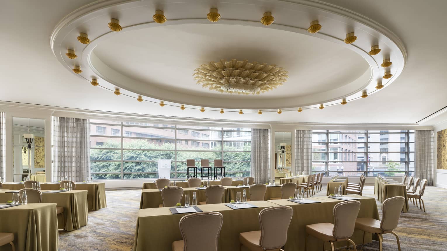 A large meeting room with large tables with beige chairs and table cloths, a large yellow chandelier hangs from the ceiing.