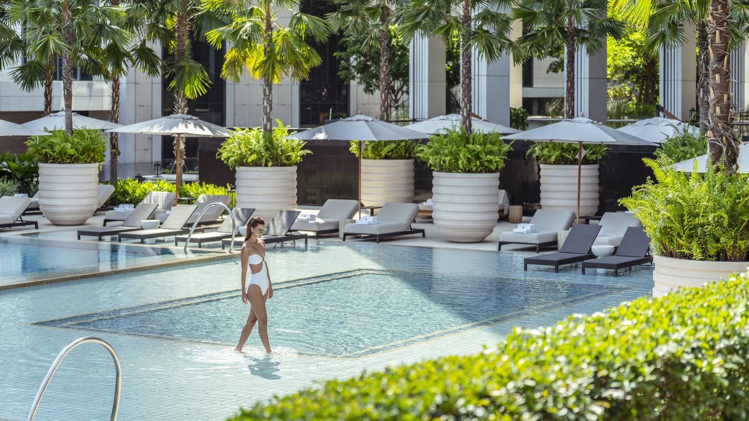 Woman in white bathing suit wades in outdoor pool surrounded by lounge chairs with umbrellas and palm trees