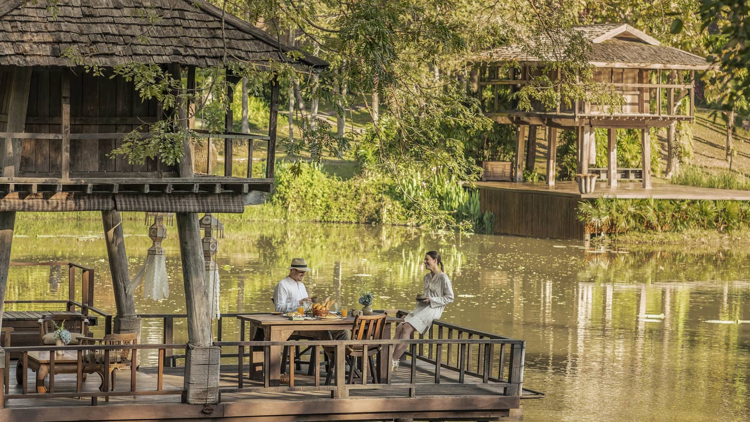 Two guests at a rustic table, on a platform in one of two wood huts on stilts perched atop a calm river bordered by greenery.