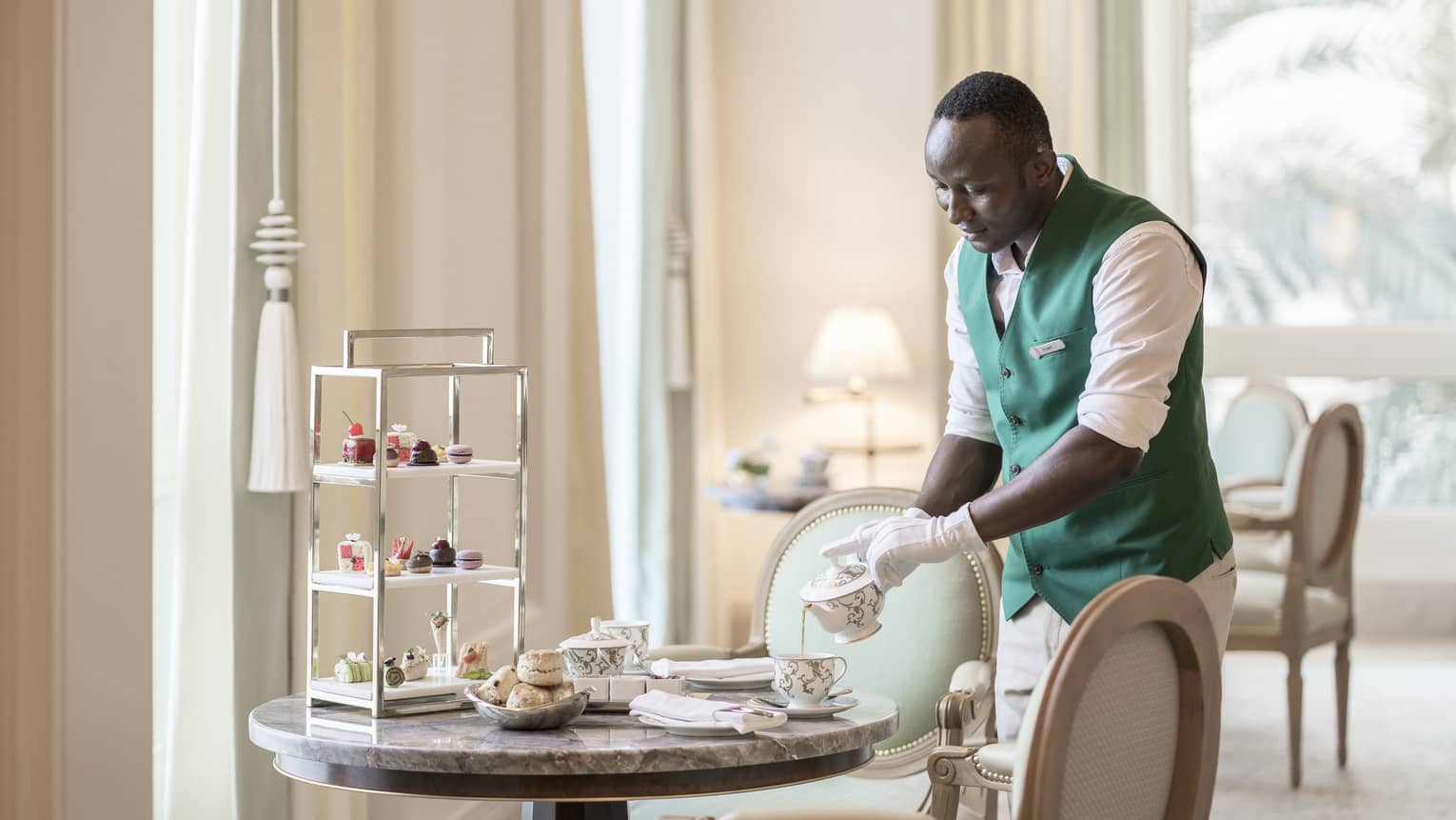 Four Seasons team member wearing a white shirt and green vest pours a cup of tea on a table set for Afternoon Tea