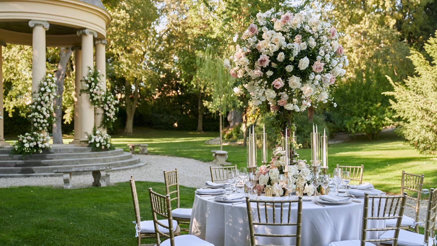 White formally set wedding reception table in garden with large white topiary and gazebo in backdrop