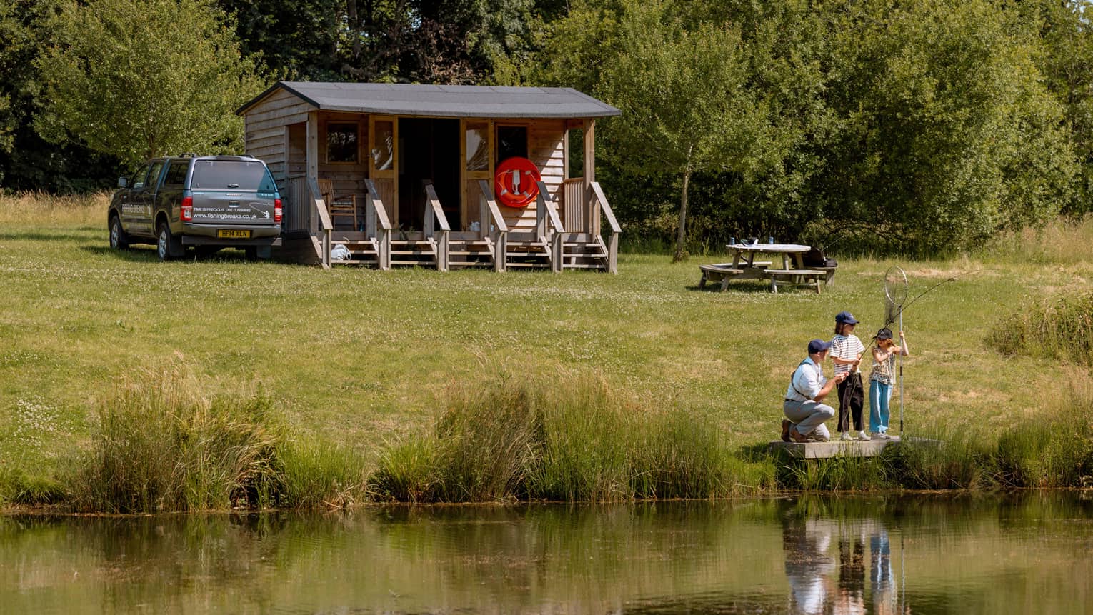 Three people fishing on small platform by lake with cabin-like building and picnic table in backdrop