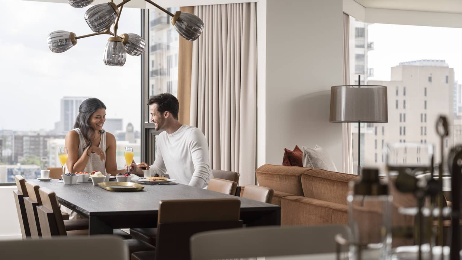 A couple is eating inside a well lit room, the city can be seen through the window behind them.