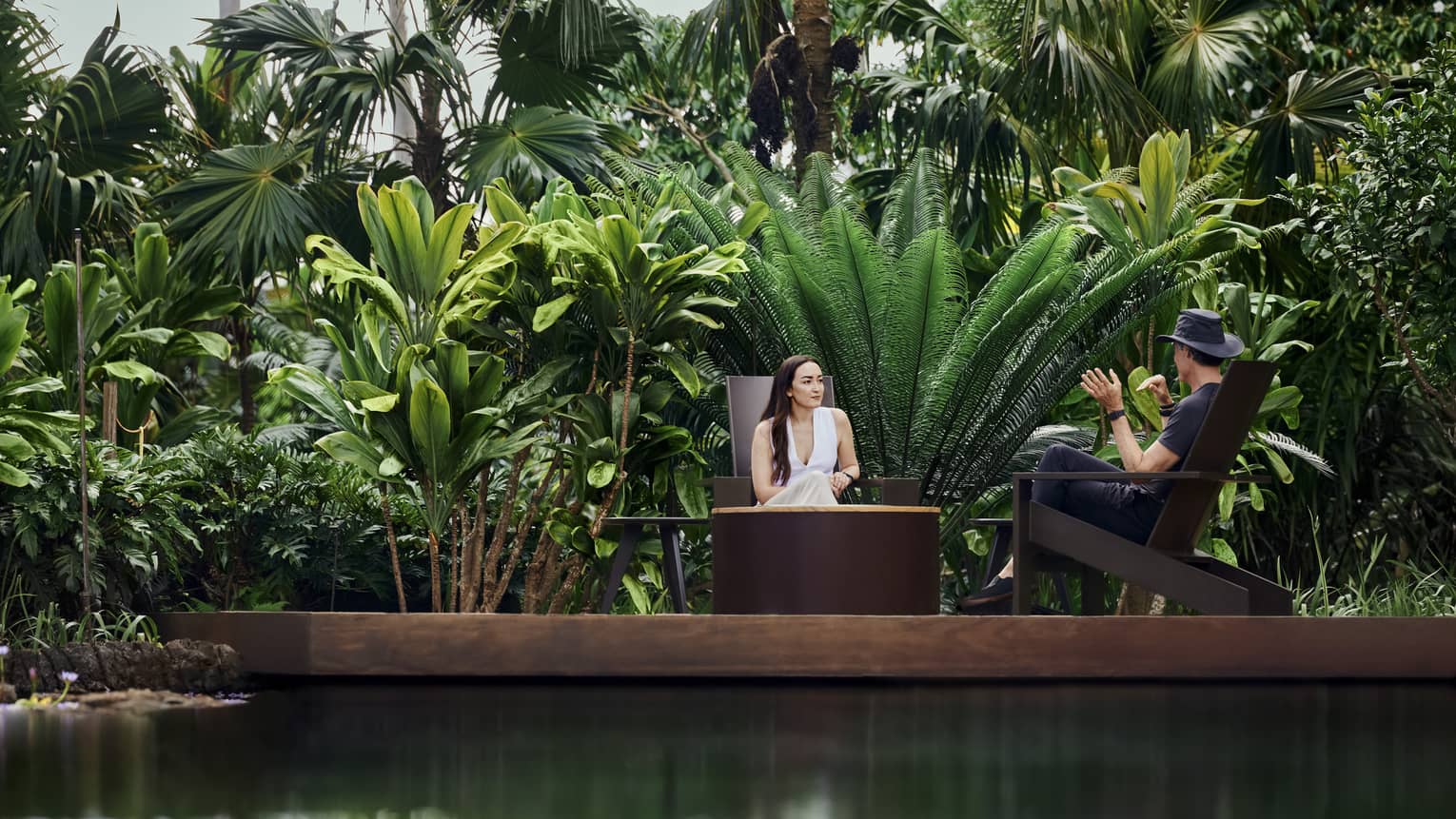 Surrounded by abundant tropical plants, two guests sit facing each atop a wooden platform over tranquil, glistening water.