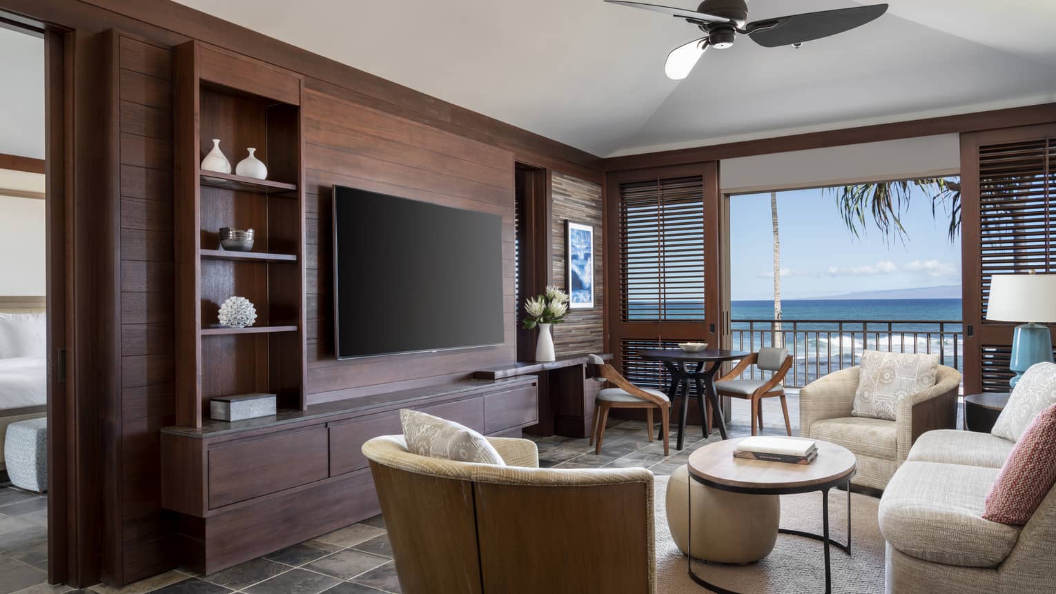 A suite's living room, with off-white sofa, two arm chairs, table, hanging flat-screen TV, walk-out balcony