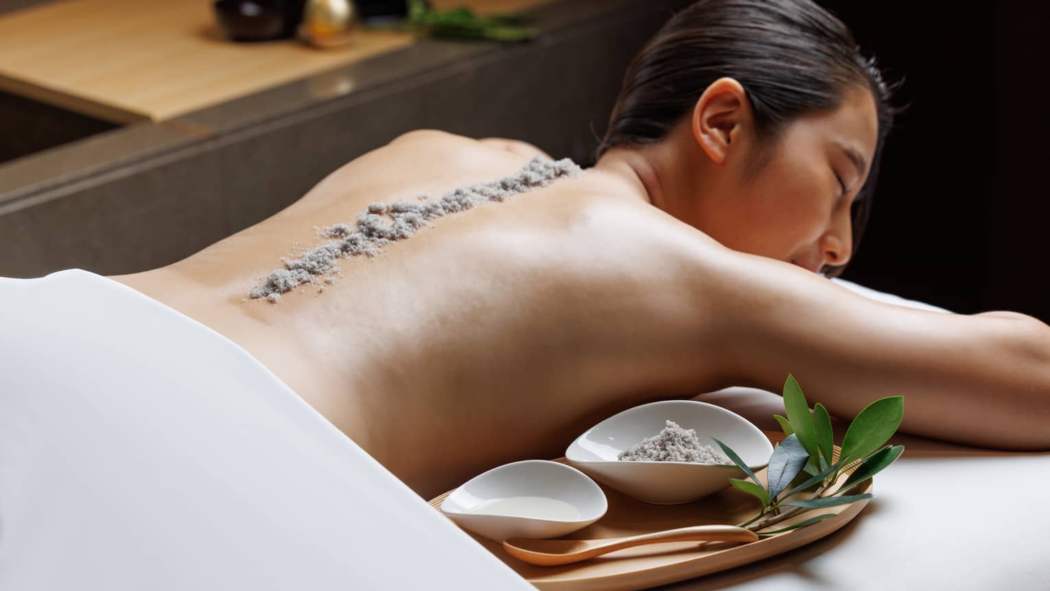 Female guest lies facedown on spa treatment table with line of exfoliating sand running the length of her spine