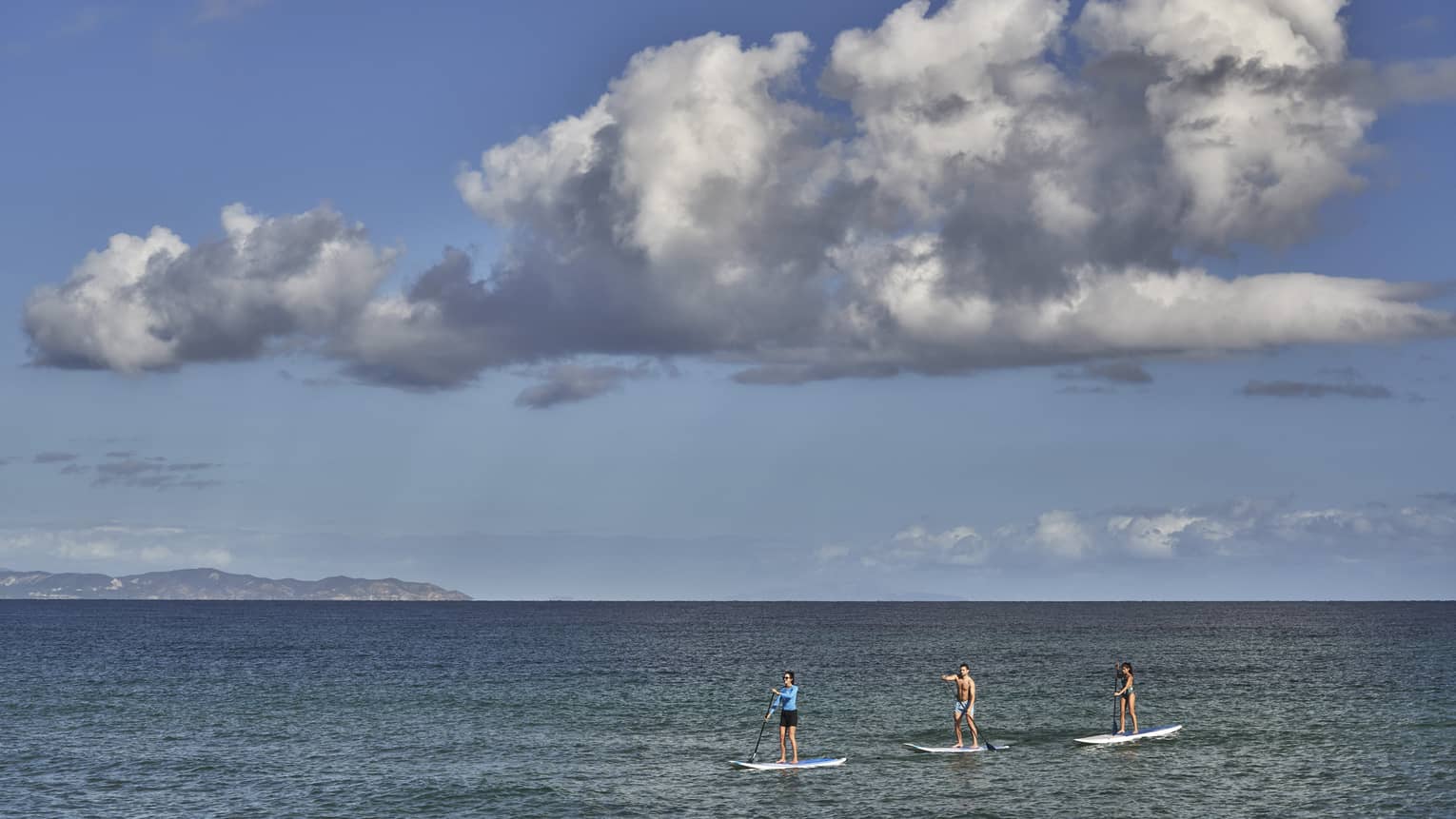 Three people on stand-up paddleboards glide on open, calm water under an expanse of blue sky and a large dramatic cloud.