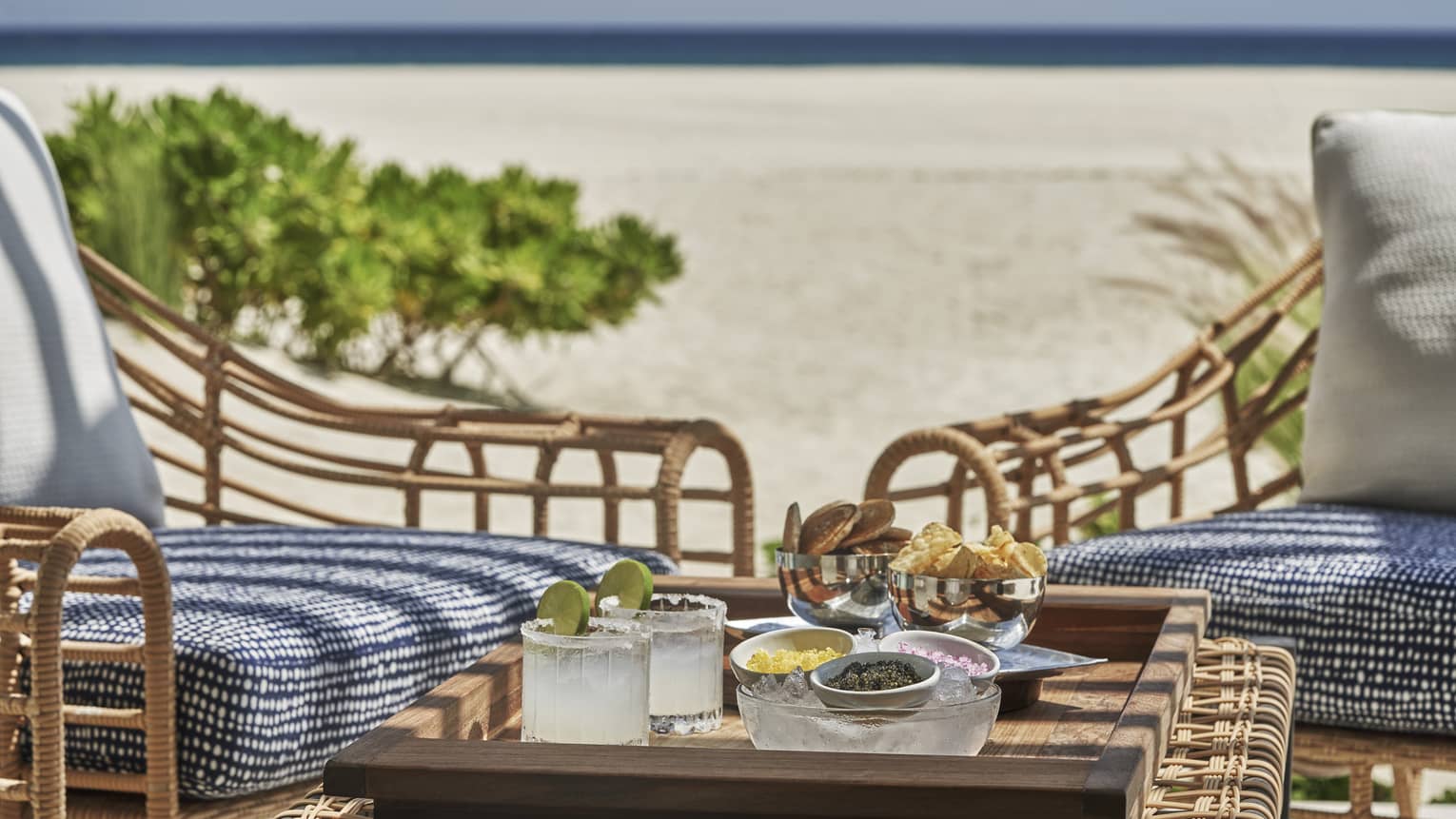 Two cushioned chairs outside with a small table with two drinks, chips and salsa on the beach.