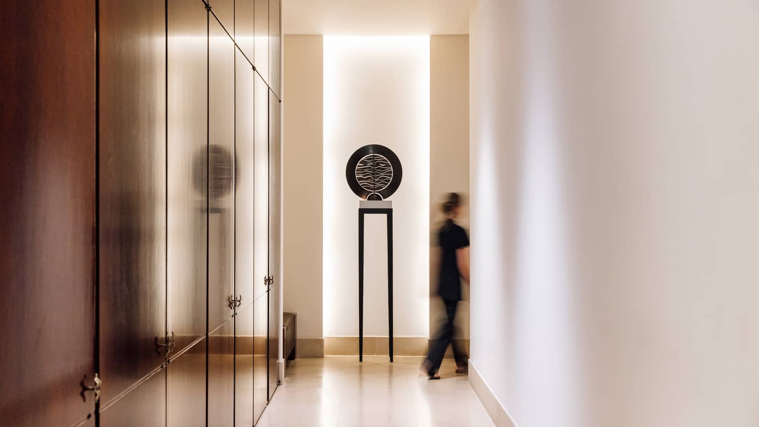 Spa hallway with art piece at the end and a spa therapist dressed in black going through a doorway beside it