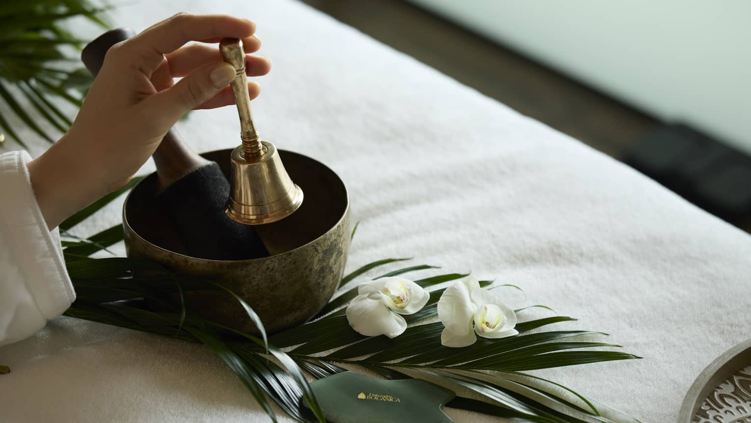 Left hand holding a brass bell over a singing bowl on a spa table