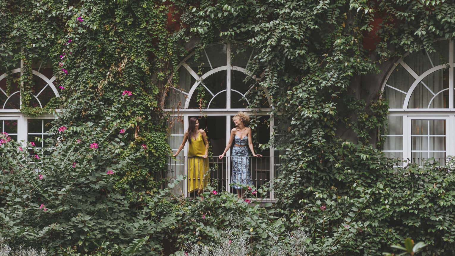 Two women stand on outdoor terrace; lush greenery covers the building and grounds