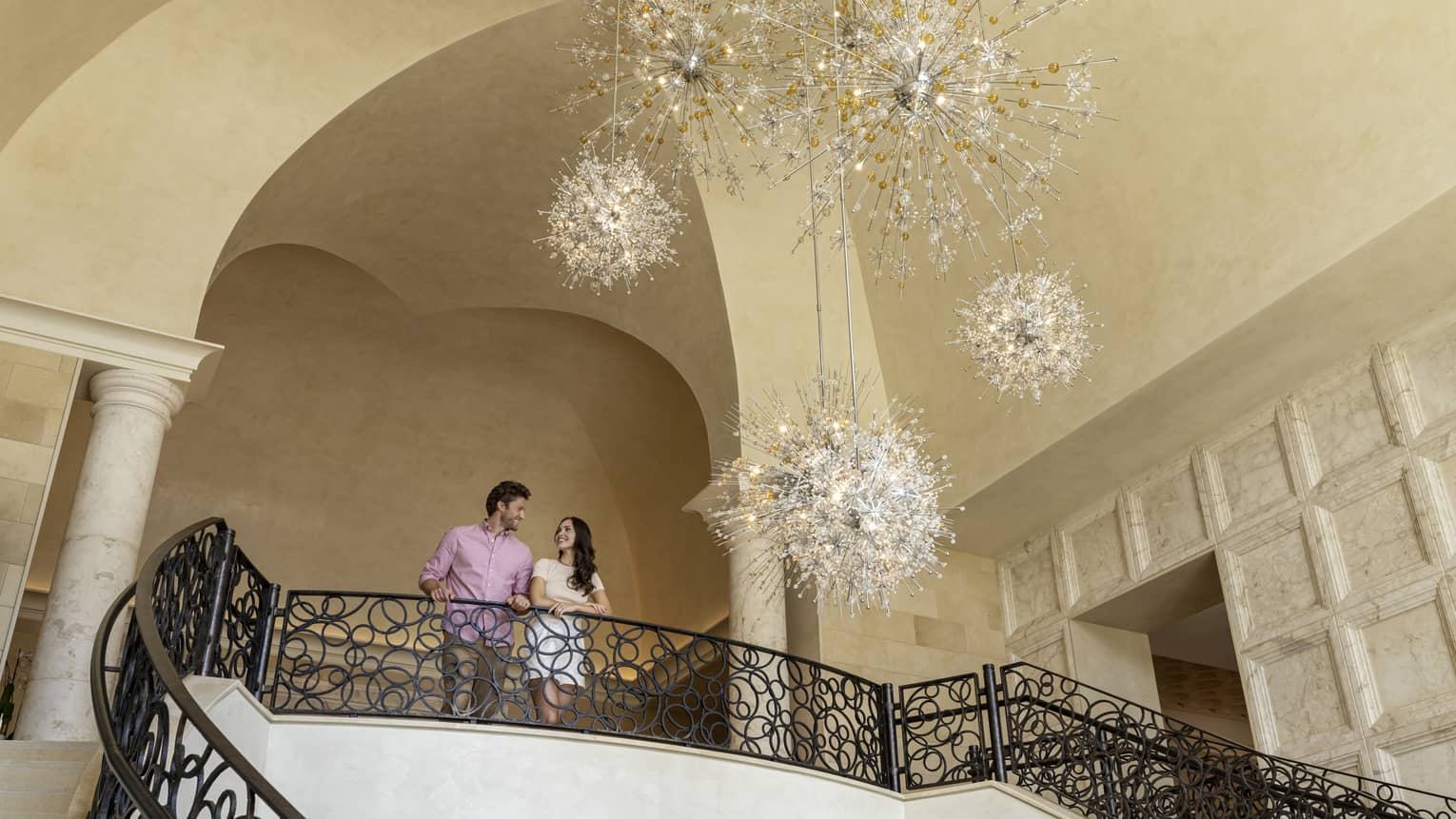 A man and woman standing at the top of a stairway with an open space looking at a large chandelier.