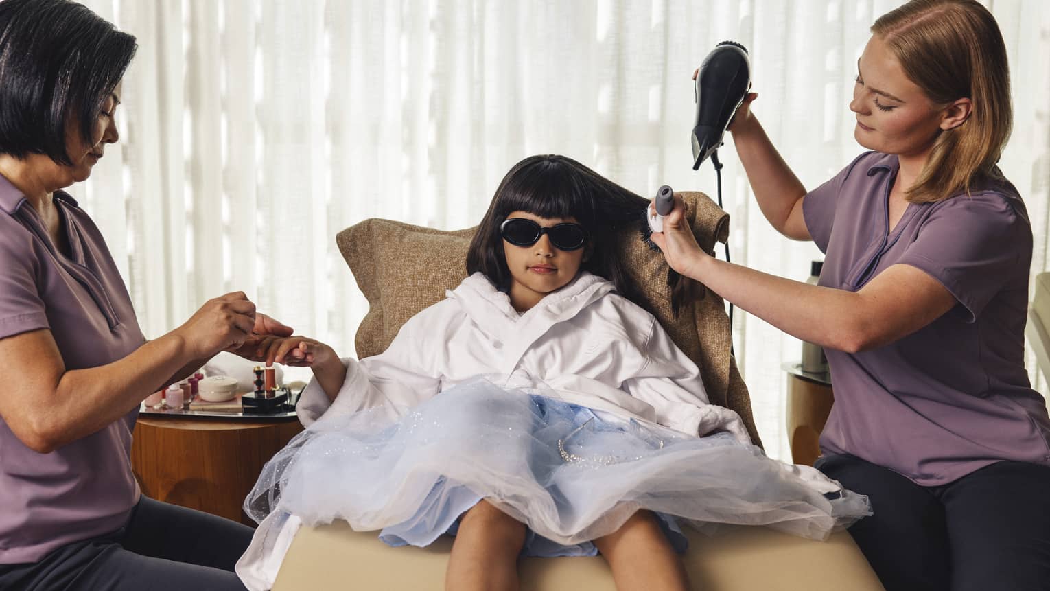 Little girl with dark brown hair and bangs lounges on a beige seat wearing a light-blue princess dress, a white robe and sunglasses as one spa attendant blow dries her hair and another paints her nails