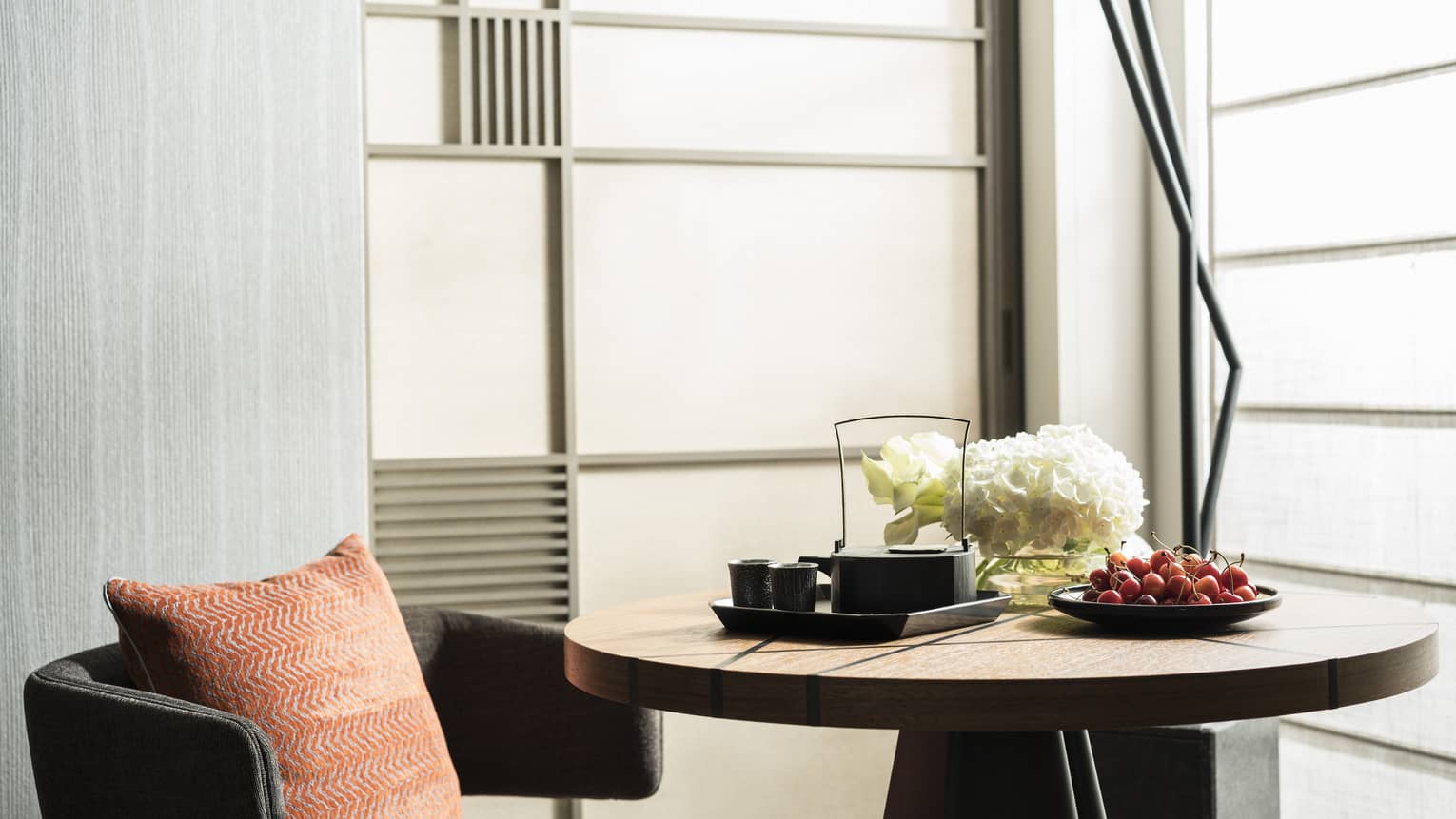 Cozy seating area with a round wooden table set with a tea tray, cherries and white flowers, featuring an orange cushion